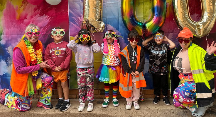photo of 5 kinder students and 2 staff members posing in front of a colourful banner